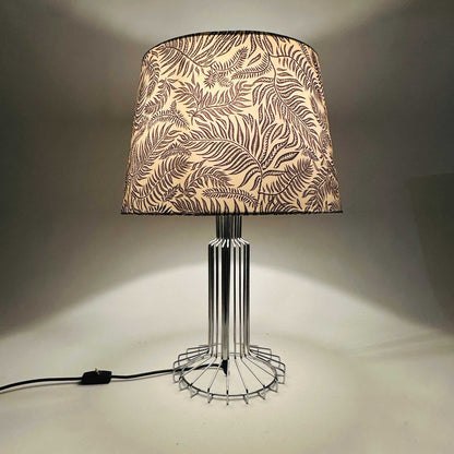 Eingeschaltete Tischlampe mit chromfarbenem Drahtfuß und weißem Stoffschirm mit schwarzem Blattdekor der 1970er