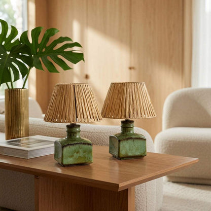 Zwei olivgrüne Keramikleuchten mit Rattanschirmen auf einem Holztisch im hellen Wohnraum